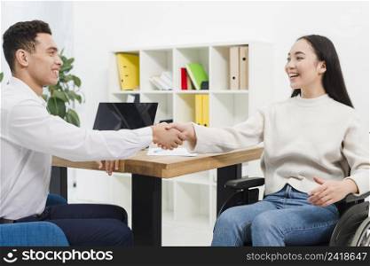 smiling young businesswoman sitting wheelchair shaking hands with businessman office