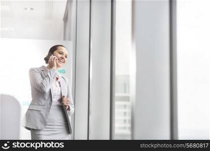 Smiling young businesswoman talking on cell phone near office window