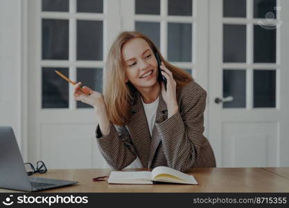 Smiling young businesswoman talks on phone writing notes, sitting at office desk. Female professional secretary personal assistant holding smartphone answers mobile call consulting client at workplace. Smiling female manager secretary talks on phone, answers mobile call, consulting client at workplace