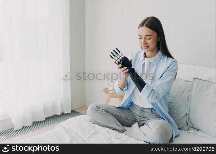 Smiling young caucasian disabled girl touching her artificial arm, sitting on bed at home. Pleased female customizing bionic prosthetic hand. High tech care about persons with disability concept.. Disabled girl touches bionic prosthetic arm at home. High tech care about persons with disability