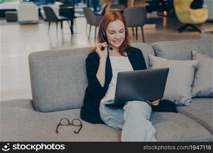 Smiling young caucasian red haired business woman working remotely online sitting on sofa in cafe with laptop, speaking with customer through wireless earphones headset, holding microphone with hand. Smiling young caucasian red haired businesswoman in headset working remotely online in cafe