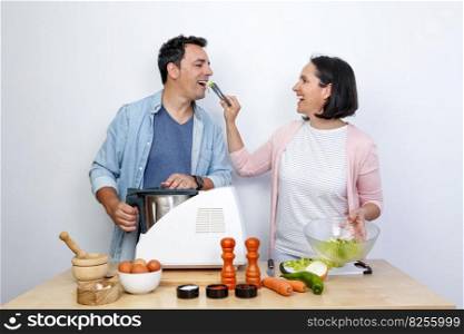 smiling young couple cooking at a table with ingredients. The woman gives the man salad with kitchen tongs.