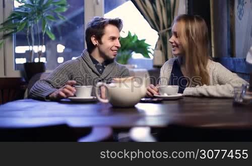Smiling young couple in love sitting at cafe table and drinking hot cup of tea while hanging out and talking at the restaurant. Two lovers enjoying time together and drinking tea in cozy coffee shop while communicating with each other.