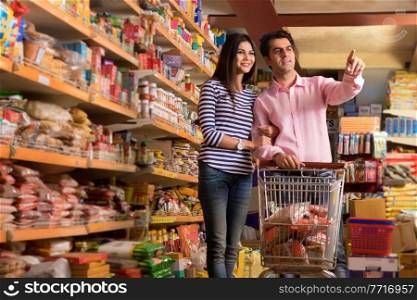 Smiling young couple looking at products at a super market holding a shopping cart while purchasing groceries.