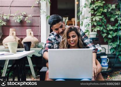 Smiling young couple sitting embraced in the steps of their wooden house with a laptop