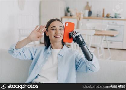 Smiling young disabled girl greeting friend, waving hand, answering video call, holding smartphone, using bionic robotic prosthetic arm sitting on couch at home. Lifestyle of people with disabilities.. Smiling disabled girl answering video call, holding smartphone, using bionic robotic prosthetic arm