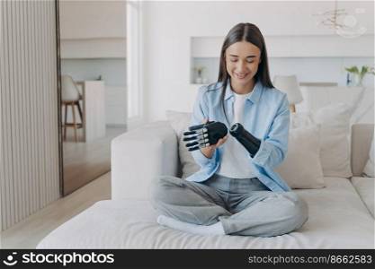 Smiling young disabled girl puts together her bionic prosthetic arm, installing artificial limb, sitting on couch at home. Young woman happy with using high tech myoelectric prosthesis.. Smiling young disabled girl puts together her bionic prosthetic arm, sitting on couch at home