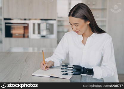 Smiling young disabled girl writing notes, holding notebook by bionic prosthetic hand, sitting at table. Satisfied pretty woman with disability writes to-do list in notepad at home.. Smiling young disabled girl writing notes, holding notebook by bionic prosthetic hand at table