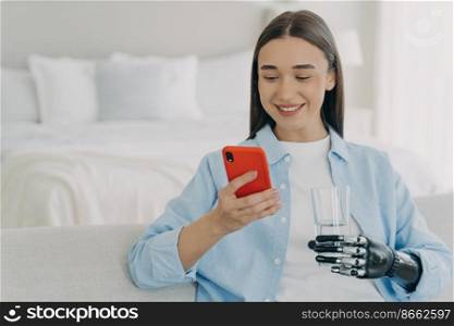 Smiling young disabled woman holding smartphone and glass of pure water. Female with bionic prosthetic arm using mobile phone apps, chatting in social networks, shopping online at home.. Disabled girl with bionic prosthetic arm using mobile phone apps at home, holding glass of water