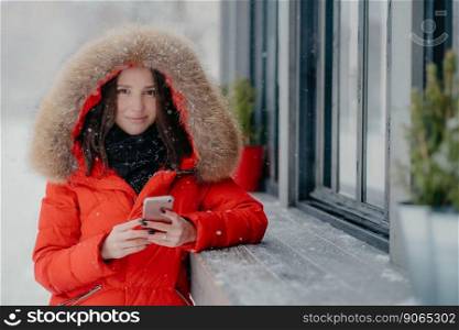 Smiling young European female in red coat, has black manicure, uses modern electronic gadget for communication in internet with friends, likes winter weather, sends photo in social networks.