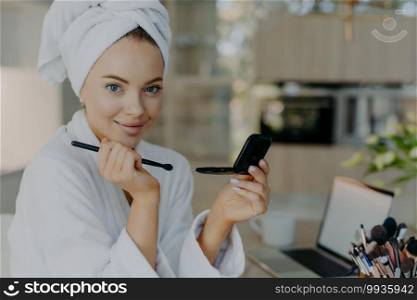 Smiling young European woman with blue eyes applies blush or powder on cheeks holds cosmetic brush puts daily makeup wears bathrobe and wrapped towel poses against modern apartment interior.