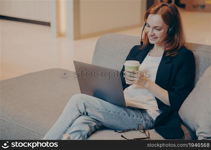 Smiling young female call center agent in wireless headset and laptop, communicates with clients of financial company, provides online consultations, while sitting on sofa in teahouse with coffee cup. Smiling young female call center agent in wireless headset and laptop sitting on sofa in cafe