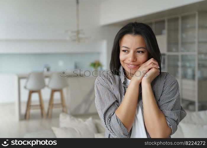 Smiling young female homeowner or tenant feels happiness and cosiness, standing at home. Happy brunette woman folded hands near cheek, satisfied new house. Mortgage, real estate sale, relocation.. Female homeowner or tenant feels happiness at new home. Advertisement of mortgage, real estate sale