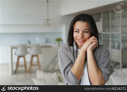 Smiling young female homeowner or tenant feels happiness and cosiness, standing at home. Happy brunette woman folded hands near cheek, satisfied new house. Mortgage, real estate sale, relocation.. Female homeowner or tenant feels happiness at new home. Advertisement of mortgage, real estate sale