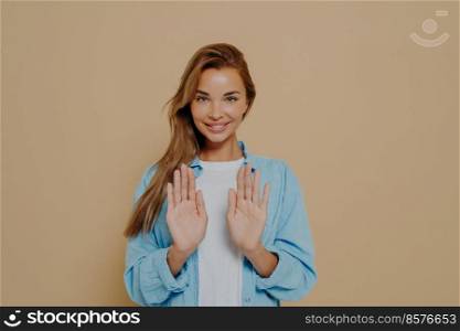Smiling young female model with stright long hair wearing oversized blue shirt and white tshirt using hands to show stop sign while standing over beige background in studio. Positive women emotions. Smiling young female model with stright long hair wearing oversized blue shirt