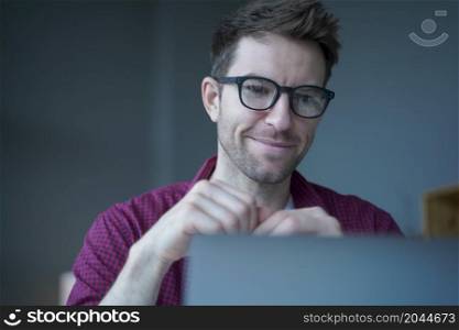 Smiling young German man freelancer in eyewear looking at marketing research report on laptop screen, analyzing statistical data and developing growth strategy, Austrian guy working remotely from home. Smiling young German man freelancer in eyewear working remotely online on laptop from home office