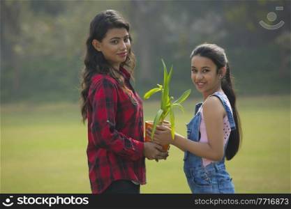 Smiling young girl giving a plant in a flower pot to her mother outdoors.