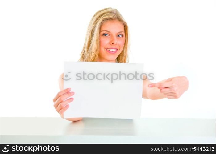 Smiling young girl sitting at table and pointing on blank paper&#xA;