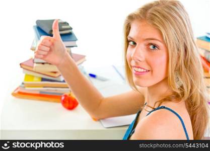 Smiling young girl sitting at table with books and showing thumbs up gesture&#xA;