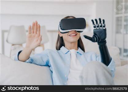 Smiling young girl wearing virtual reality glasses learning to use bionic prosthetic arm after limb loss. Disabled woman in vr goggles testing artificial hand, myoelectric prosthesis.. Smiling young girl wearing virtual reality glasses learning to use high tech bionic prosthetic arm