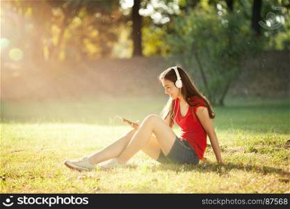 Smiling Young Girl with Pleasure Listening to Music on Headphones on the Mobil Phones Sitting on the Meadow in the fine Summer Day