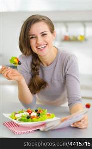 Smiling young housewife eating fresh salad and reading magazine in kitchen