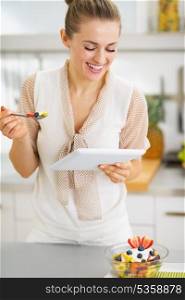 Smiling young housewife eating fruits salad and using tablet pc