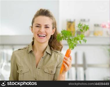 Smiling young housewife holding carrot in kitchen