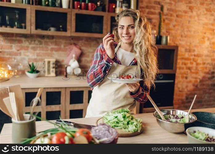 Smiling young housewife in apron prepares salad, kitchen interior on background. Female cook making healthy vegetarian food, vegetables preparation. Smiling young housewife in apron prepares salad