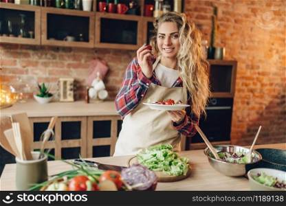Smiling young housewife in apron prepares salad, kitchen interior on background. Female cook making healthy vegetarian food, vegetables preparation. Smiling young housewife in apron prepares salad