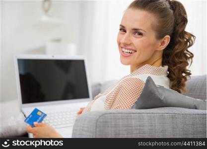 Smiling young housewife with laptop and credit card sitting on couch