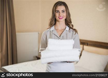 smiling young maid holding white stack towels standing front bed