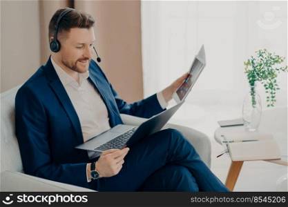 Smiling young male boss in suit using headset to make video call with employees, looking at laptop screen and discussing report while sitting in armchair in living room of his apartment. Executive manager reviewing documents during online meeting at home