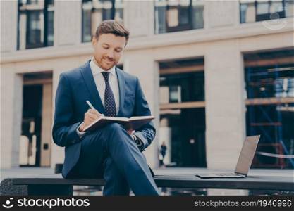 Smiling young male enterpruer working onling on laptop outside, taking notes in notebook while sitting on bench in front of office building with crossed legs, using laptop for distand work. Smiling young man dressed in blue suit taking notes in notebook while spending time outdoors