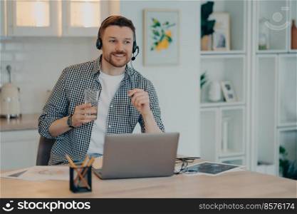 Smiling young male freelancer in headset taking part in online meeting or web conference on laptop, holding glass of water and pen while working at home. Remote job concept. Happy man in casual clothes going to make notes in notebook while studying at home