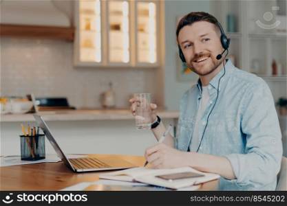 Smiling young man freelancer or entrepreneur working from home and using headset with microphone during an online conference, holding glass of water and looking aside. Remote job concept. Bearded freelancer making notes in notebook while sitting at his workplace at home