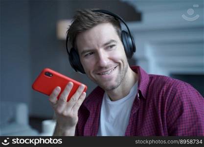 Smiling young man German language tutor in headphones speaking with student on cellphone, joyful austrian guy recording voice message for collegue, using speakerphone while remotely online from home. Smiling young German man in headphones recording voice message for collegue, using speakerphone