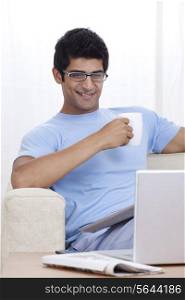 Smiling young man having coffee while looking at laptop