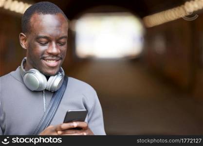 Smiling Young Man In Urban Setting Checks Social Media And Messages On Mobile Phone Whilst Wearing Wireless Headphones