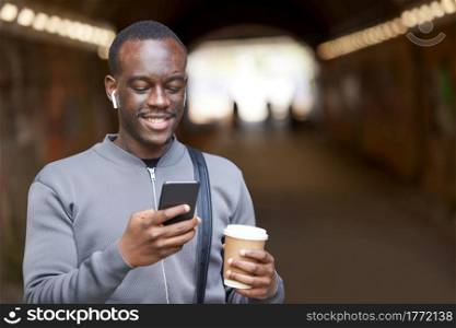 Smiling Young Man In Urban Setting Checks Social Media And Messages On Mobile Phone Whilst Wearing Wireless Earphones