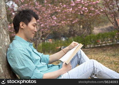 Smiling young man leaning on a tree and enjoying his book, outdoors in a park