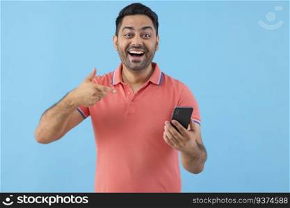 Smiling young man looking at camera and pointing at Smartphone holding in his hand 