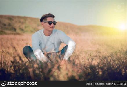 Smiling young man sitting on the grass, smiling young man in eyeglasses sitting on the field, concept of happiness in the field