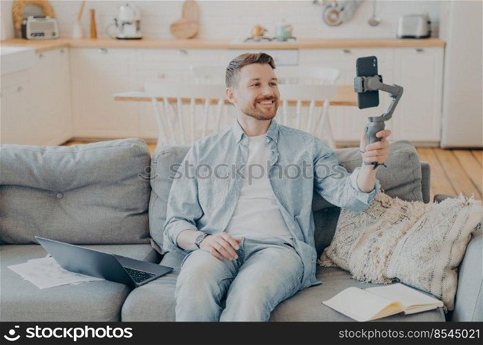 Smiling young man talking to his friend and showing him his apartment while in video call, holding gimbal with phone, sitting on couch beside laptop, open note book and papers. Young man in video call with friend while sitting on couch