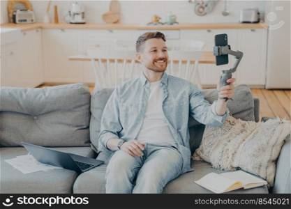 Smiling young man talking to his friend and showing him his apartment while in video call, holding gimbal with phone, sitting on couch beside laptop, open note book and papers. Young man in video call with friend while sitting on couch