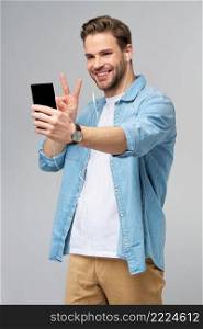 Smiling young man wearing jeans shirt taking selfie photo on smartphone or making video call standing over grey studio background.. Smiling young man wearing jeans shirt taking selfie photo on smartphone or making video call standing over grey studio background