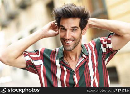 Smiling young man with dark hair and modern hairstyle wearing casual clothes in urban background.. Smiling young man with dark hair and modern hairstyle wearing casual clothes outdoors