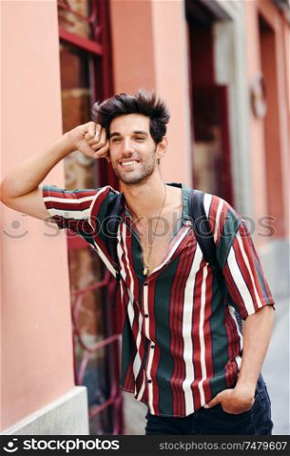 Smiling young man with dark hair and modern hairstyle wearing casual clothes in urban background.. Smiling young man with dark hair and modern hairstyle wearing casual clothes outdoors