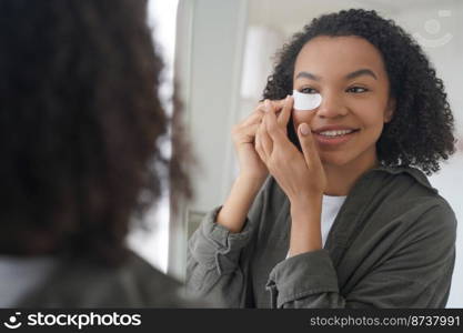 Smiling young mixed race girl applying under eye patches on face, looking in mirror. Beautiful teen lady with afro hairstyle, enjoying daily skincare, beauty routine. Modern cosmetics advertising.. Smiling young mixed ace girl applying under eye patches looking in mirror. Skincare, beauty routine