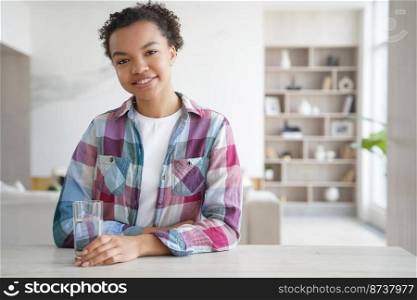 Smiling young mixed race girl sitting at table with glass of clean mineral water at home. Happy biracial lady following healthy lifestyle, drinking pure seltzer. Diet, habit for for wellness.. Happy mixed race girl sitting with glass of pure mineral water at home. Diet, healthy lifestyle